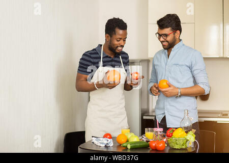 Deux beaux mâles joyeux amis debout dans la cuisine de fruits dans les mains, parler, faire du jus d'orange et de pamplemousse, la préparation des aliments sains t Banque D'Images