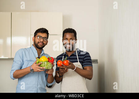 Portrait de deux chefs masculins debout dans cuisine légère holding fresh légumes bio à sourire à la caméra. Multi Ethnic friends preparing salad Banque D'Images