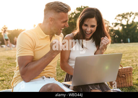 Portrait d'heureux couple homme et femme criant et célébrons les succès tout en utilisant un ordinateur portable en parc d'été Banque D'Images