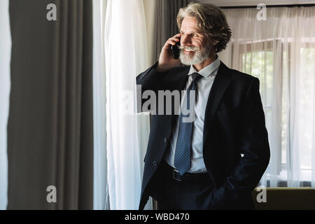 Image d'adultes joyeux businessman wearing costume noir parler sur smartphone en étant debout près de fenêtre dans hotel apartment Banque D'Images