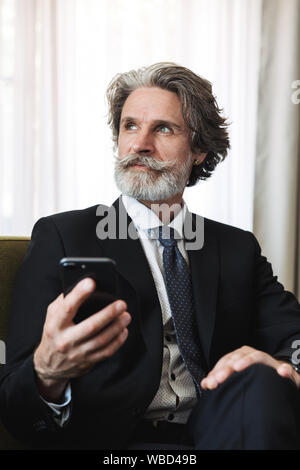 Photo d'une grave concentré aux cheveux gris senior business man sitting in chair à l'intérieur à l'aide d'un téléphone mobile. Banque D'Images