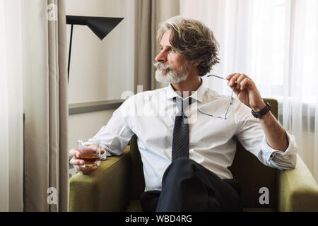 Photo d'un aux cheveux gris happy business man sitting in chair à l'intérieur à la maison l'alcool holding glasses dans la main. Banque D'Images