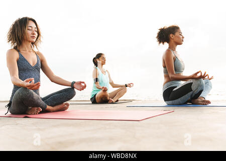 Photo d'ambiance les sportives multiethniques en survêtements de méditer et de faire des gestes zen tout en étant assis sur un tapis de yoga par mer à matin Banque D'Images