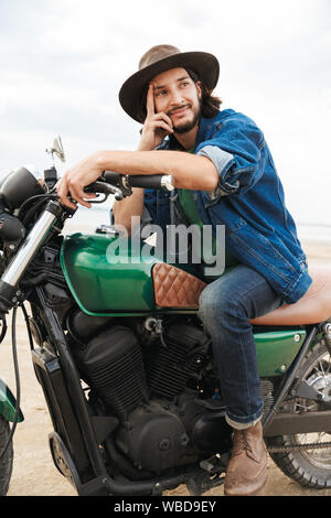 Handsome smiling young man wearing tenue décontractée assis sur une moto à la plage, de porter un chapeau Banque D'Images