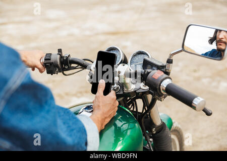 Close up of a man holding guidon et à l'aide de téléphone mobile tout en étant assis sur une moto à la plage Banque D'Images