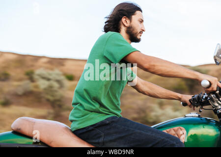 Vue latérale d'un beau jeune homme portant tenue décontractée circonscription une moto à la plage Banque D'Images
