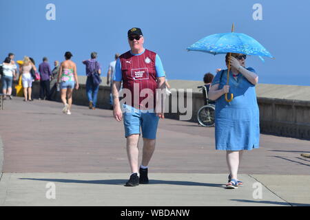 Homme dans un maillot de football, femme avec parapluie bleu, Scarborough, North Yorkshire, Royaume-Uni, 26th août 2019, Météo: Chaud et ensoleillé août vacances à la banque lundi après-midi et les records de température devraient être brisés dans l'est de l'Angleterre pour la fin des vacances d'été. Les gens affluent vers le front de mer et la plage de plusieurs kilomètres autour pour s'amuser au soleil dans cette ville traditionnelle de bord de mer anglais sur la côte nord-est. Banque D'Images