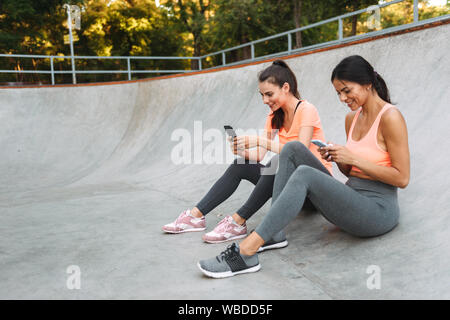 Droit des jeunes femmes dans les vêtements de sport fitness smiling, téléphones cellulaires, tandis qu'assis sur des terrain de sport Banque D'Images