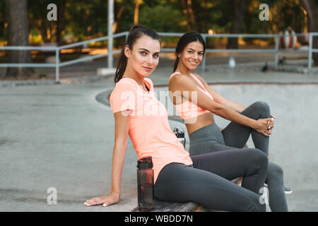 Image de l'athletic smiling women in sportswear assis avec des bouteilles d'eau sur un terrain de sport en béton Banque D'Images