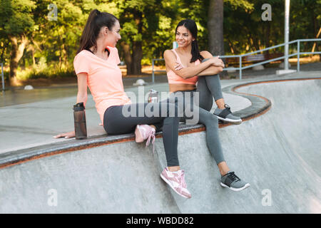 Image of beautiful women in sportswear assis et parlant avec des bouteilles d'eau sur un terrain de sport en béton Banque D'Images