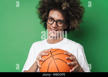 Gros plan Portrait of smiling caucasian man with afro hairstyle holding basketball et looking at camera isolated over green background Banque D'Images