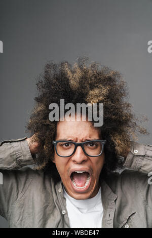 Gros plan Photo de jeune homme a souligné avec afro hairstyle attrapant sa tête et crier et isolé sur fond gris Banque D'Images