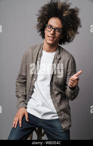 Photo de heureux homme non rasé avec coiffure afro assis sur une chaise et doigt d'avance sur fond gris isolé Banque D'Images