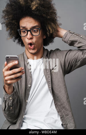 Gros plan Photo de jeune homme choqué avec coiffure afro à la téléphonie cellulaire et à toucher sa tête sur fond gris isolé Banque D'Images