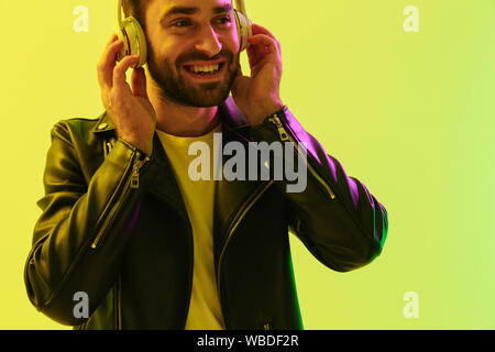 Photo de sourire heureux beau jeune homme élégant posant sur fond vert clair isolé avec mur de néons led habillé en veste en cuir Banque D'Images