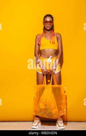 Image pleine longueur d'african american woman wearing sunglasses debout et portant le sac en plastique sur fond jaune isolé Banque D'Images