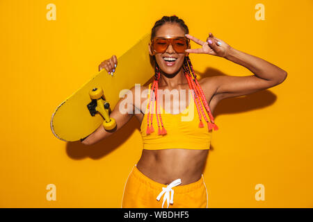 Droit de funny african american woman wearing sunglasses holding skateboard et gestes isolés signe de paix sur fond jaune Banque D'Images