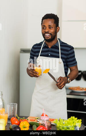 Photo de beau jeune homme à la peau foncée eyewear holding glass avec le jus d'orange fraîchement pressé, profitant de boisson savoureuse à la lumière à la cuisine Banque D'Images