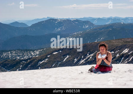 Une fille est assise dans la neige dans une robe d'été sur le pic enneigé de la montagne et la neige tient dans ses mains. La jeune fille sourit. Dans le contexte de la ma Banque D'Images