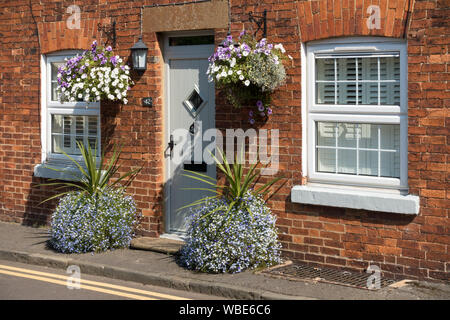 Vieux chalet moderne en brique rouge avec double vitrage PVC et porte avant avec de jolies fleurs en pots et paniers suspendus, Uppingham, Rutland, UK. Banque D'Images