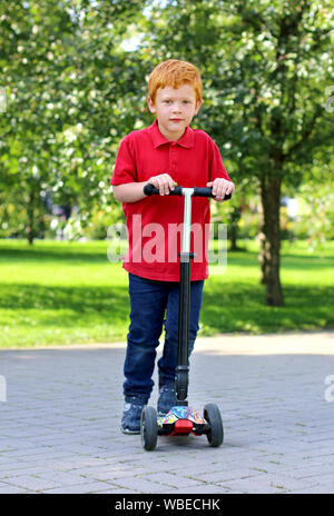 Enfant apprendre à conduire un scooter dans un parc de la ville aux beaux jours d'été. Joyeux petit garçon aux cheveux gingembre posant avec un scooter. Loisirs et plein air active Banque D'Images