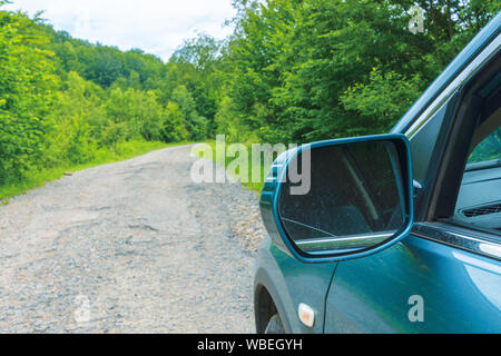 Vus sur l'ancienne route forestière. bleu cyan véhicule stationné sur le chemin d'asphalte fissuré en amont entre les arbres. vue depuis la porte du conducteur et rétroviseur latéral. perdu d Banque D'Images