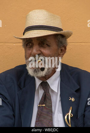 Trinidad, Cuba, 26 nov., 2017 - vieil homme est assis sur un banc de fumer un cigare, contre un mur jaune Banque D'Images