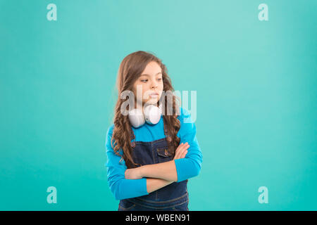 Hipster kid. Petit hipster en gardant les bras croisés sur fond bleu. Hipster adorable avec de longs cheveux bouclés portant des casques modernes. Mignon petit enfant dans un style branché. Banque D'Images