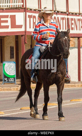 Woman wearing colorful shirt, jeans et chapeau et avec de la bière peut dans la main à cheval cheval brun foncé le long de la rue principale de la ville à Quorn en Australie du Sud Banque D'Images