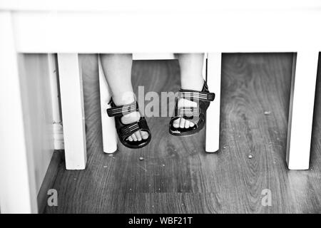 Jeune enfant assis à la petite table blanche. Pieds en sandales sous vide, suspendu au-dessus du sol. Ne peut pas toucher le sol. L'alimentation a chuté sur le sol. Banque D'Images