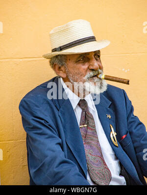 Trinidad, Cuba, 26 nov., 2017 - vieil homme est assis sur un banc de fumer un cigare, contre un mur jaune Banque D'Images