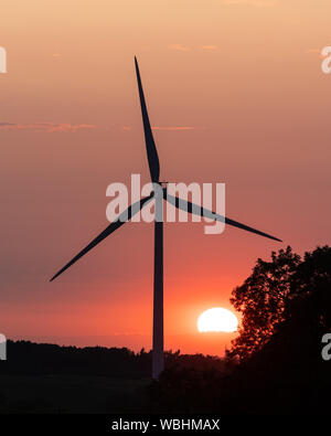 Crick, Northamptonshire, Royaume-Uni: Le soleil, partiellement obscurci par les nuages, se fixe dans un ciel orange derrière une éolienne et un arbre silhouetté. Banque D'Images