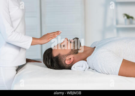 Portrait de guérisseur mettant les mains au-dessus de la tête de bel homme sur la table de massage Banque D'Images