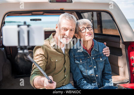 Smiling senior couple sitting on car, embrassant et en tenant selfies Banque D'Images