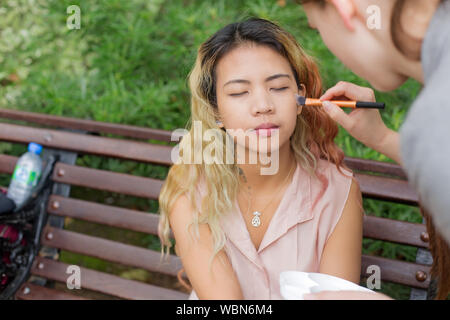 Femme modèle asiatique assis sur un banc de parc tout en appliquant le maquillage sur ses joues Banque D'Images