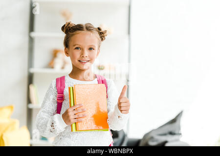 Happy schoolgirl holding books et showing thumb up à la maison Banque D'Images