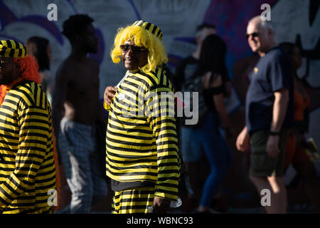 Un homme à l'Notting Hill Carnival portant un costume noir et jaune et porte une perruque jaune tout en marchant à travers la foule. Banque D'Images