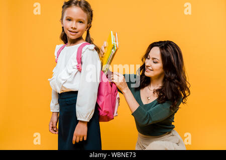 Cheerful mère mettre des livres dans un sac à dos de lycéenne isolé sur orange Banque D'Images