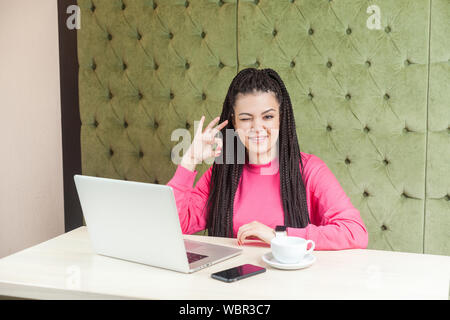 Portrait de jeune femme drôle positive avec des dreadlocks hairstyle dans noir chemisier rose assis dans un café et montrant ok signe avec la main, avec un clin, à la Banque D'Images