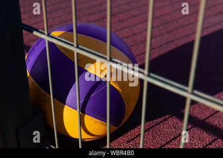 Un seul ballon allongé sur le sol rouge après le match. Équipement de volley-ball perdu. Terrain de sport au coucher du soleil avec une clôture métallique. Banque D'Images