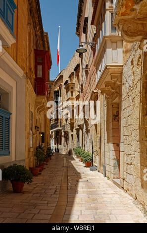 City scene ; rue piétonne étroite, de vieux bâtiments en pierre calcaire, soleil, ombre, balcons, plantes en pot, Malte drapeau, site de l'UNESCO ; Europe ; Birgu, Vittorios Banque D'Images