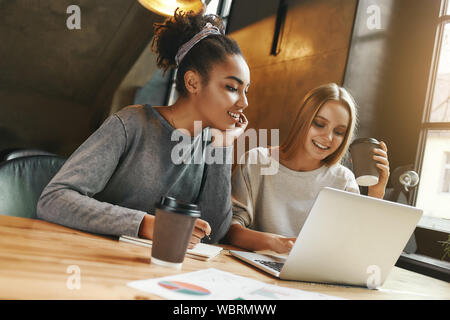 Portrait de deux jeunes femmes multiraciales amis en surfant l'internet ensemble sur un ordinateur portable et des notes qu'ils siègent dans une cafétéria prendre une tasse de café. Plan horizontal. Angle néerlandais Banque D'Images