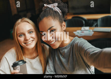 Portrait de deux jeunes femmes multiraciales en souriant tout en prenant l'aide de selfies smartphone comme ils sont assis dans une cafétéria. Plan horizontal Banque D'Images