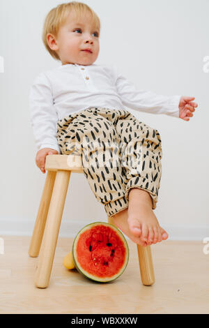 Peu de tout-petit garçon assis sur la selle. Demi tranche de pastèque rouge est en dessous. Banque D'Images