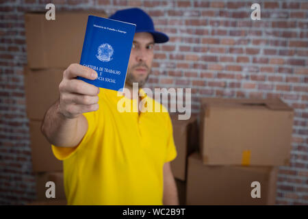 Mailman brésilien tenant son carnet de travail sécurité sociale brésilienne document, dans un dépôt avec un beaucoup de boîtes. Banque D'Images