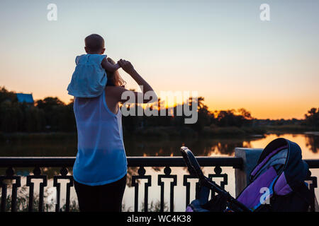 Baby Girl sitting sur les épaules de sa mère et admire le paysage. Jeune mère marcher avec kid par la rivière au coucher du soleil. Temps de la famille Banque D'Images