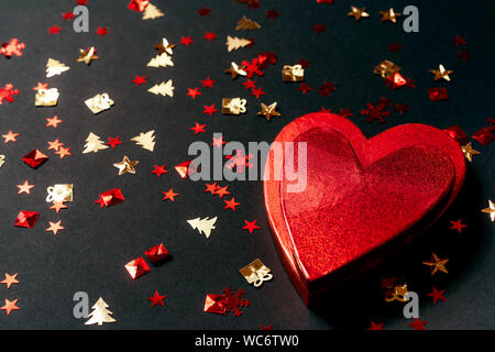 Red Heart-shaped gift box et festives confetti doré sur fond noir Banque D'Images