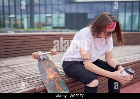 Portrait de sports brunette avec le verre dans ses mains, assis sur le banc en bois près de roulettes sur journée d'été en ville Banque D'Images