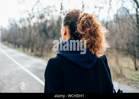 Vue arrière jeune femme avec queue de coiffure et cheveux bouclés portant manteau avec capuche hood en avant marche sur route vide en stationnement à journée d'hiver. Banque D'Images