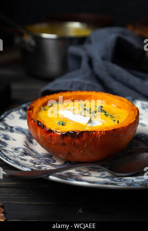 Soupe de potiron crémeux cuit servi dans une petite citrouille de moitié sur une plaque en bois vintage sur fond rustique, orientation verticale Banque D'Images